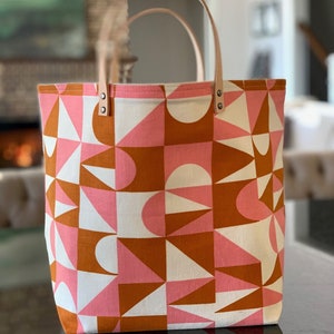 May include: A large tote bag with a geometric pattern in shades of pink, white, and brown. The bag has leather handles and a zipper closure.