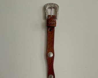 May include: A brown leather belt with silver-toned metal accents and a decorative buckle. The belt has a wavy design and features five round metal buttons.