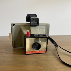May include: A gray Polaroid Land Camera, the Big Swinger 3000, with a red button and a black lens. The camera has a black strap.