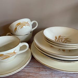 May include: Cream-colored vintage dishware set with gold trim and wheat stalk design. Includes teacups, saucers, bowls, and plates. The set is arranged on a wooden surface, showcasing a classic and elegant aesthetic.