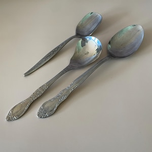 Peut inclure: Trois cuillères en argent vintage avec des motifs décoratifs sur les manches. Les cuillères sont de tailles et de formes différentes, dont une cuillère de service. Elles sont disposées sur une surface claire, reflétant la lumière ambiante.