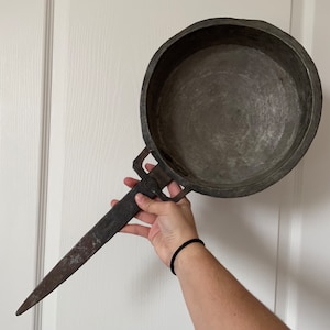 Op de afbeelding: Een vintage, ronde metalen pan met een lange, dunne steel. De pan is gemaakt van donker metaal en heeft een versleten, rustieke uitstraling. De steel is met een metalen lus aan de pan bevestigd.