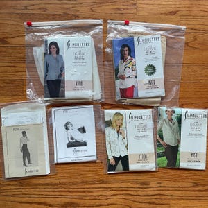 May include: Several vintage sewing pattern packets in clear plastic bags are arranged on a wooden surface. The packets feature images of women modeling blouses and jackets. The brand name "Silhouettes" is visible on the packets, along with pattern numbers.