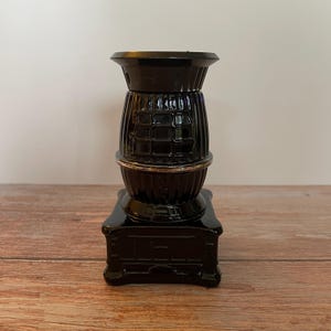 Op de afbeelding: Een zwarte decoratieve kandelaar met een uniek ontwerp. De kandelaar heeft een vierkante basis, een cilindrisch middengedeelte met een rasterpatroon en een uitlopende bovenkant. De houder is gemaakt van een glanzend materiaal en staat op een houten oppervlak.