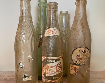 May include: A collection of five vintage glass soda bottles. The bottles are various shapes and sizes, with faded labels and signs of age. One bottle features a Pepsi-Cola label, and another has a faded cartoon graphic.