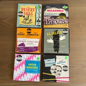 May include: A collection of six vintage 45 RPM record sleeves. The sleeves feature a variety of musical artists and genres, including country, Broadway, and classical. The sleeves are in good condition and have a retro look.