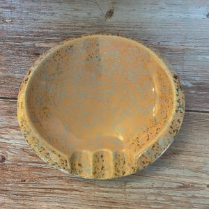 May include: A vintage ashtray with a peach-coloured interior and a speckled, light blue and gold rim. The ashtray has three grooves for resting cigarettes. The ceramic ashtray is round.