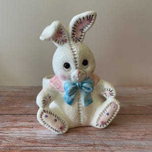 Può includere: Statuetta di coniglio in ceramica bianca con fiocco blu e dettagli rosa. Il coniglio ha grandi occhi neri, interno delle orecchie rosa e dettagli cuciti. È seduto con le gambe distese.