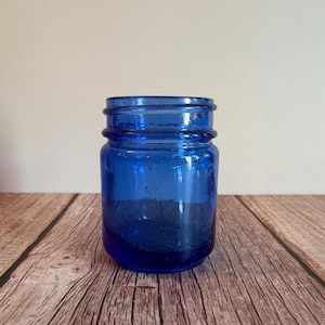 May include: A small, cobalt blue glass jar with a wide mouth and a thick, ridged rim. The jar is cylindrical with a slightly rounded base. The glass has a textured appearance, and the jar sits on a wooden surface.