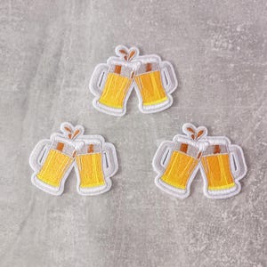 Könnte beinhalten: Drei gestickte Aufnäher mit jeweils zwei anstoßenden Bierkrügen. Die Krüge sind mit gelbem Bier gefüllt und haben weiße Umrisse. Über jedem Paar befindet sich ein kleiner brauner Spritzer. Auf grauem Hintergrund.