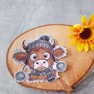 Könnte beinhalten: Ein skurriles Patch mit einer braunen Kuh, die eine graue Strickmütze und einen Schal trägt und einen traurigen Gesichtsausdruck hat. Das Patch befindet sich auf einer Holzscheibe, neben einer gelben Sonnenblume.