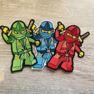 Könnte beinhalten: Drei gestickte Stoffaufnäher mit stilisierten Ninja-Figuren in Grün, Blau und Rot. Jede Figur trägt eine passende Maske und Uniform mit goldfarbenen Akzenten und hält zwei Schwerter hinter dem Rücken, umrandet von schwarzen Linien.