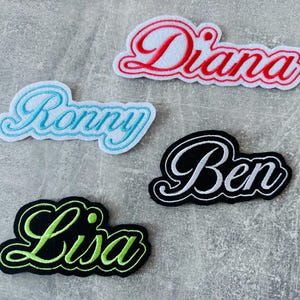 Könnte beinhalten: Vier gestickte Namensaufnäher: "Diana" in Rot und Weiß, "Ronny" in Blau und Weiß, "Ben" in Schwarz und Weiß und "Lisa" in Schwarz und Grün. Jeder Aufnäher hat eine Schreibschrift mit kontrastierendem Rand, auf grauem Hintergrund.