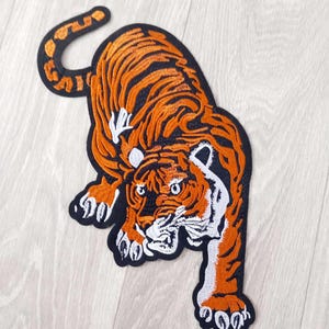 Könnte beinhalten: Ein gestickter Aufnäher, der einen springenden Tiger darstellt. Der Tiger ist orange mit schwarzen Streifen und weißen Akzenten an Gesicht und Pfoten. Das gesamte Motiv ist mit einem dicken schwarzen Rand versehen, der eine klare Form bildet.