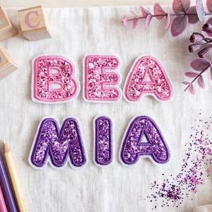 Könnte beinhalten: Ein Set aus sechs bestickten Buchstabenaufnähern, die den Namen BEA MIA bilden. Die Buchstaben B, E und A sind rosa mit glitzernden Texturen, während die Buchstaben M, I und A lila mit glitzernden Texturen sind. Jeder Buchstabe hat einen weißen Rand.