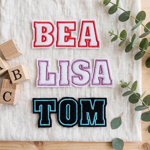 Könnte beinhalten: Drei bestickte Namensaufnäher auf einem weißen Stoffhintergrund. Der oberste Aufnäher zeigt BEA in Rot, der mittlere LISA in Lila und der untere TOM in Schwarz. Jeder Buchstabe ist in einer fetten Schriftart im College-Stil gehalten.