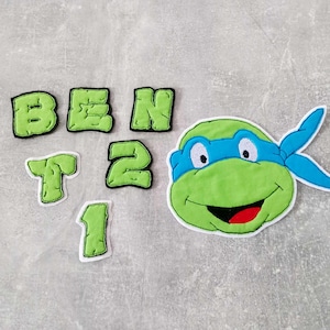 Könnte beinhalten: Grüne Filzbuchstaben, die "BEN" buchstabieren, und die Zahlen "1", "2" und "T" mit einer grünen und blauen Filzfigur der Teenage Mutant Ninja Turtles.