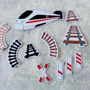 Könnte beinhalten: Ein Set aus Filzapplikationen mit einem weißen, schwarzen und roten Hochgeschwindigkeitszug, verschiedenen gebogenen und geraden braunen und schwarzen Gleissegmenten, einem weiß-roten Bahnübergangsschild und drei weiß-rot gestreiften Schildern.