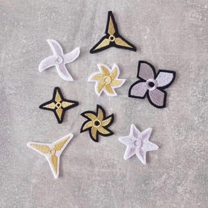 Könnte beinhalten: Eine Sammlung von acht gestickten Aufnähern in verschiedenen Stern- und Blumenformen. Die Aufnäher sind in Gold, Weiß und Grau mit schwarzen Umrandungen gehalten. Jedes Stück weist ein zentrales kreisförmiges Detail und geometrische Muster auf.