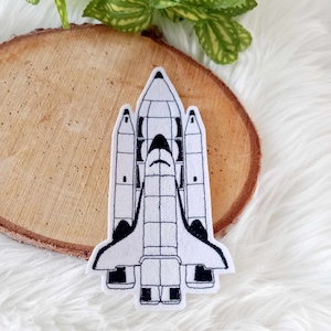 Könnte beinhalten: Ein gestickter Aufnäher aus weißem Filz, der ein Raumschiff mit schwarzen Umrissen zeigt. Das Raumschiff ist nach oben gerichtet, mit der Nase zum oberen Bildrand.