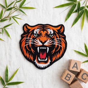 Könnte beinhalten: Ein gestickter Aufnäher mit einem Tigerkopf mit orangefarbenem, schwarzem und weißem Fell. Der Tiger hat ein offenes Maul mit rotem Zahnfleisch, weißen Zähnen und grünen Augen. Das Design ist auf schwarzem Hintergrund mit detaillierten Stichen.