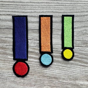 Könnte beinhalten: Drei gestickte Ausrufezeichen-Patches in Blau, Orange und Grün, jeweils mit schwarzem Rand. Der blaue Patch hat einen roten Kreis, der orangefarbene einen hellblauen Kreis und der grüne einen gelben Kreis. Auf einer Holzmaserung.