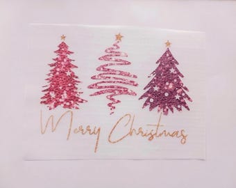 Pode incluir: Um guardanapo de papel retangular branco com três árvores de Natal estilizadas em tons de rosa e roxo com texturas brilhantes. Cada árvore é rematada com uma pequena estrela dourada. O texto Merry Christmas está impresso em cursivo dourado.