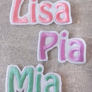 Könnte beinhalten: Drei gestickte Filzpatches mit den Namen Lisa, Pia und Mia. Die Patches sind rosa, lila und grün und haben einen weißen Umriss.