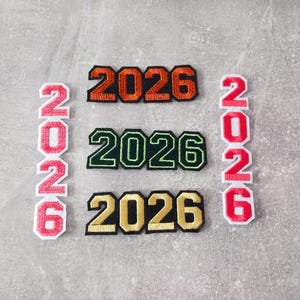 Könnte beinhalten: Gestickte Aufnäher mit der Jahreszahl "2026" in verschiedenen Farbkombinationen. Die Aufnäher umfassen Orange, Grün und Gold mit schwarzen Umrissen sowie vertikale rote Aufnäher mit weißen Umrissen.