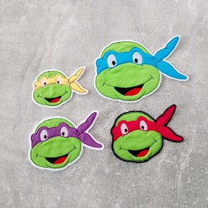 Könnte beinhalten: Vier gestickte Teenage Mutant Ninja Turtles Gesichts-Patches. Jeder Patch zeigt eine Maske in einer anderen Farbe: blau, gelb, lila und rot.