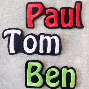 Könnte beinhalten: Drei bestickte Namensaufnäher mit den Namen Paul, Tom und Ben. Der obere Aufnäher hat rote Buchstaben auf schwarzem Grund, der mittlere Aufnäher hat weiße Buchstaben auf schwarzem Grund und der untere Aufnäher hat hellgrüne Buchstaben auf Schwarz.