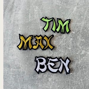 Könnte beinhalten: Drei bestickte Namensaufnäher mit Graffiti-Schriftzug und schwarzem Rand. Der oberste Aufnäher zeigt TIM in Limettengrün, der mittlere MAX in Goldgelb und der untere Aufnäher zeigt BEN in weißem Garn auf einem schwarzen Hintergrund.