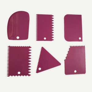 May include: Set of six pink plastic cake decorating tools. The tools have different shapes and edges, including a rounded spatula, a straight spatula with a serrated edge, a triangle spatula, and a scalloped spatula.