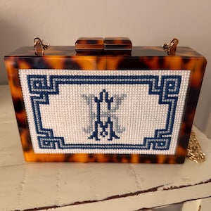 May include: A rectangular box clutch with a brown and black tortoiseshell pattern frame. The center features a white needlepoint panel with a blue Greek key border and a central monogram featuring the letters K and M in blue and light gray thread.