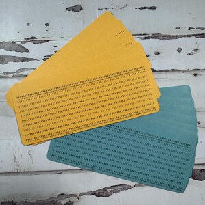 May include: A stack of rectangular yellow and teal paper data processing cards. Each card features a grid of printed numbers arranged in horizontal rows, with columns numbered from zero to eighty across the top edge of the card surface.