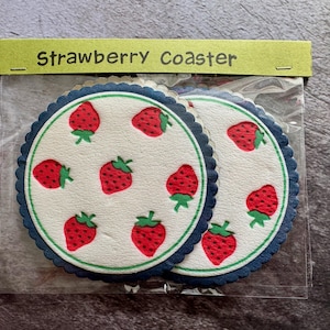 Può includere: Due sottobicchieri rotondi con sfondo bianco, con un motivo di fragole rosse con steli verdi. I sottobicchieri hanno un bordo smerlato blu scuro. La confezione recita "Strawberry Coaster".