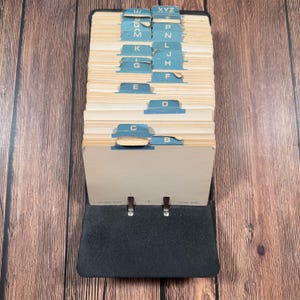 May include: A black rotary card file organizer featuring a series of cream-colored index cards with blue alphabetical tabs ranging from B to XYZ. The base is black with two metal rings holding the cards in place, and the text reads ROLLER NOTE.