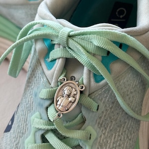 May include: A light mint green athletic sneaker with matching laces and a silver-toned oval metal charm featuring an embossed figure of Saint Sebastian attached to the center of the lacing system. The shoe has bright turquoise accents.