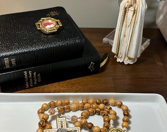 May include: A wooden rosary with a silver crucifix and a gold medallion, displayed on a white tray. The rosary features light brown beads. A statue of the Virgin Mary and a closed black book are also visible.