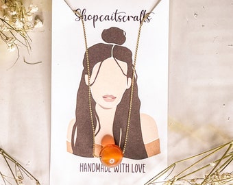 May include: A gold chain necklace with an orange carnelian stone pendant. The necklace is displayed on a white card with the text "Shopcaitscrafts" and "Handmade with Love".
