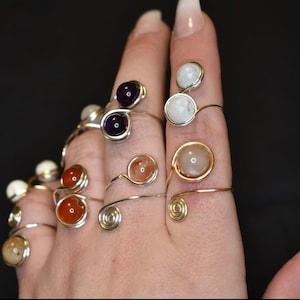 May include: A hand wearing multiple wire wrapped rings with various colored gemstones. The rings are made with silver and gold wire and feature amethyst, carnelian, and white stones.