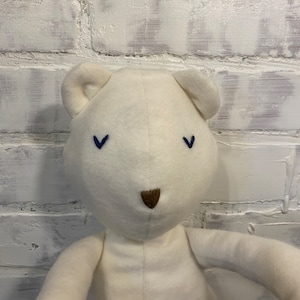 May include: A white plush teddy bear with blue embroidered eyes and a brown nose. The bear is sitting on a white brick wall background.