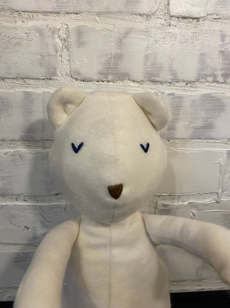 May include: A white plush teddy bear with blue embroidered eyes and a brown nose. The bear is sitting on a white brick wall background.