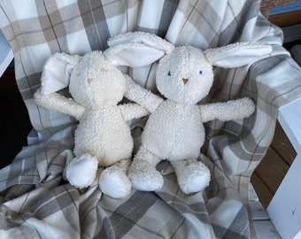 May include: Two white plush bunny toys with long ears are sitting on a cream and brown plaid blanket. The blanket has a fringe on the bottom.