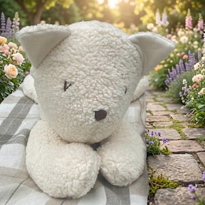 May include: A cream-colored plush toy shaped like a cat with a textured, curly fleece-like exterior. The toy features simple embroidered eyes and a small, dark brown nose. It is resting on a light-colored fabric with a grey and white plaid pattern.
