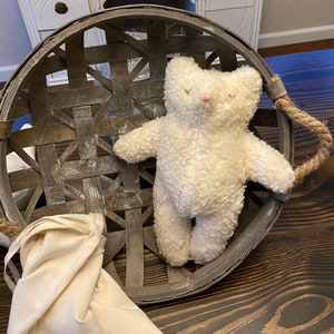 May include: A white plush teddy bear with a pink nose sits inside a woven basket with a brown wooden handle.