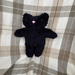 May include: A small, black teddy bear with a fluffy texture. The bear has a pink nose and brown embroidered eyes and ears. It is set against a plaid background in shades of white, beige, and brown.
