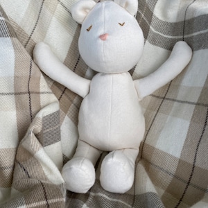 May include: A white plush teddy bear with closed eyes and a pink nose, lying on a plaid blanket.