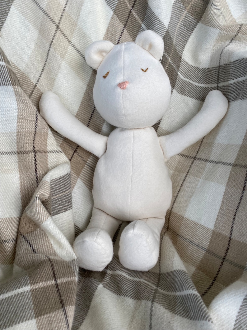 May include: A white plush teddy bear with closed eyes and a pink nose, lying on a plaid blanket.