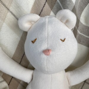 May include: White plush toy bear with embroidered eyes and a pink nose. The bear is lying on a plaid blanket.
